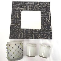 Wall mirror with 'Laugh' text design on the frame placed alongside three candle holders on a white surface.