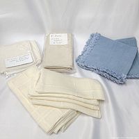 All napkins arranged on white surface showing varied styles and colors including beige hemstitched cocktail napkins, ecru linen napkins, blue tassel napkins, and eggshell napkins.