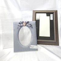 Two picture frames shown side by side on a white background. One frame is grey-brown wood with satin finish and black mat, the other light grey satin with floral/ribbon decor and oval opening.
