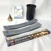 Lot overview showing brass cone incense burner and base, quartz angel wing necklace in packaging, black pillar candle holder, two soapstone incense holders, and three boxes of HEM incense sticks.