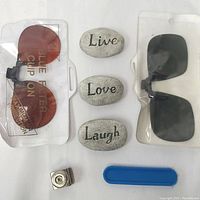 Overview of the lot including both sunglass clip-ons in plastic packaging, the three inspirational stones, the vintage metal hole puncher, and the blue plastic item.