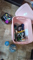 Pink plastic tote partially open showing assortment of art supplies inside including brushes, tubes of paint, spackling compound, and tools.