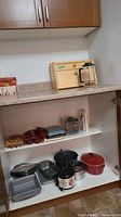 Full view inside wooden kitchen cabinet showing multiple baking pans, silicone and tin muffin trays, coffee press, small kitchen tools, and wooden bread box on countertop