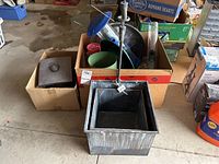 Photo showing two nested square galvanized tin planters with decorative hanging faucet tap, placed on concrete floor with some boxes and garden items in background.