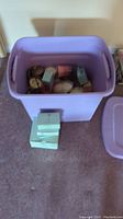Purple plastic tote filled with various candles including some in glass jars and boxes piled inside and outside the tote.