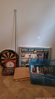 View of dartboard with clock, framed puzzle, curtain, rod, and blue tote filled with books and CDs on floor.