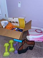 Overview photo showing box with orange waist support belt, blue Fit Hoop, and pink Fit Hoop box, along with yellow and purple hand weights and orange-black ankle support band.