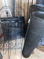 Partial rolls of black plastic garden screening along with stacked black metal garden fencing panels featuring decorative designs and fleur-de-lis finials.