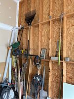 Photo of multiple long-handled tools like rakes, hoe, spade, bamboo stakes, and hose with hanger hanging on an OSB wood wall.
