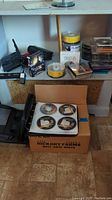 Overview photo showing various media items including boxed video games, compact disc spindles, cassette tapes, a disc carrying case with game discs, yellow and white blank media spindles on a shelf, and a cardboard box on the floor containing a black CD binder open to show four video game discs.