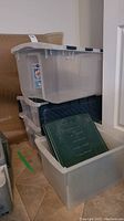 Two stacked translucent plastic storage totes with gray lids, one white tote below with a dark green lid resting inside, on tile flooring.