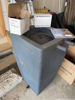 Full view of the grey resin rain barrel standing on the floor with the pump kit box placed on top and instruction materials visible.