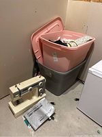 Janome sewing machine with instruction book placed on ground beside two plastic bins containing assorted fabrics.