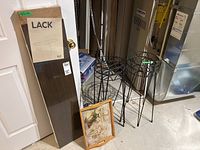 Photo showing the IKEA Lack shelf in packaging leaning against a wall. Four black metal wire plant stands arranged nearby on floor. Rectangular wooden serving tray with visible water stains placed in front of shelf and plant stands.