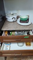 White Lancaster slow cooker on countertop with large white serving tray holding green ceramic and small glass bowls. Open drawers showing kitchen utensils and flatware.