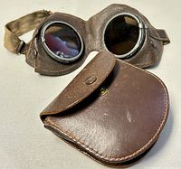 Antique leather motorcycle goggles placed in front of a brown leather carrying case with snap button flap.