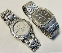 Two men's watches side by side on a light fabric surface: ESQUIRE round silver-tone watch with a link bracelet and Pulsar rectangular silver-tone watch with a black dial and link bracelet.