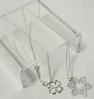 Two 925 silver necklaces displayed on a clear acrylic stand, showing chain and pendants. The four-leaf clover pendant is visible alongside the floral pendant.
