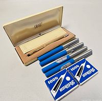 Set of four Pelikano fountain pens with chrome caps and eight blue Soffix ink cartridges, arranged next to a closed vintage Cross pen case.