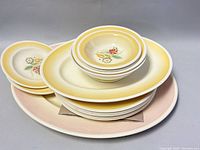 Stack of yellow-rimmed plates and bowls atop large pink oval platters, showing floral patterns and size comparison.