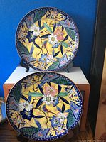 Photo of two large decorative plates with colorful floral and bird patterns, placed on display stands against a blue wall.