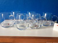 Photo showing six clear glass mugs arranged in a row on a wooden surface against a dark blue wall, highlighting their round shape and clear glass.