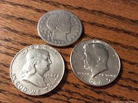 Three half dollar coins displayed on wooden surface showing front faces of 1962 Franklin, 1969 Kennedy, and 1914 Barber coin.