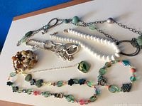 Photo showing assortment of bracelets and necklaces laid out on a surface with blue background, displaying beads, chains and clasps in various colors and materials.