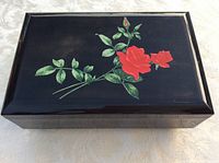 Closed rectangular jewelry box with glossy black laquered finish and red rose painting on top, displayed flat on a white textured cloth.