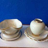 Front view of stacked and grouped Lenox porcelain pieces including bowls, shell-shaped plates, small vase with gold accents on white surface