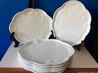 Two milk glass plates displayed upright with 6 plates stacked flat showing embossed grape and leaf design.