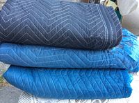 Stack of three quilted packing blankets in black, medium blue and dark blue color, showing chevron stitching detail.