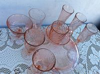 Nine pieces of pink vintage glass dinnerware arranged on lace cloth, including footed cake plate, tall glasses, and smaller dessert cups.