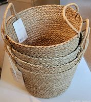 Stacked lot of four hand braided seagrass baskets showing texture, shape, and handles with brand tag visible.