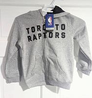 Front view of a grey hoodie for kids featuring the text 'TORONTO RAPTORS' printed in black, full zipper, and two front pockets. Hood has a contrasting black lining. NBA official tag attached.