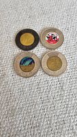 Four Canadian two-dollar coins displayed on a textured light fabric. Coin designs visible include Queen Elizabeth II, colorized 50th anniversary, Northern Lights, and Canadian beaver.