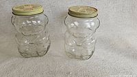 Two empty 1988 Kraft honey bear glass jars with metal lids placed on beige fabric.
