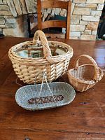 Photo showing three baskets on a wooden table: a large wicker basket with fabric lining, a small round wicker basket, and an elongated gray metal basket with decorative handle.