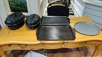Overview of five kitchen items on wooden desk: two black roasting pans with lids on left, two rectangular metal cookie sheets in center, round metal pizza pan on right.
