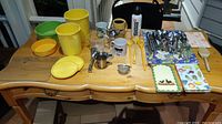 Photo showing an assortment of vintage kitchen items on wooden table including Tupperware canisters, metal spoons, mugs, and various kitchen tools