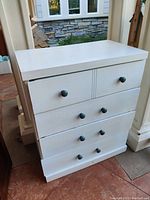 Front view of white painted wooden dresser with four drawers and visible black round knobs. Clean overall with minor wear.