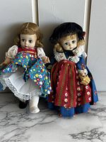 Front view of two Madame Alexander dolls standing on marble surface against white background. Left doll in white dress with blue floral apron, right doll in traditional German outfit with bonnet.