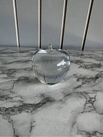 Photo of clear crystal apple-shaped paperweight, showing front side with short stem on top, placed on marble surface.