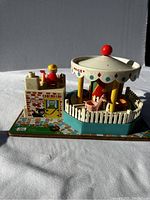 Side view showing ticket booth with Little People figure and carousel with animal seats inside white picket fence and colorful base.