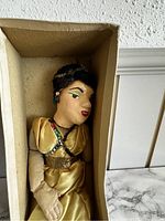 Marionette doll head and upper body detail in original box. Gold dress visible with sash and bead decoration. Face painted with black hair, green eyes, and red lips.