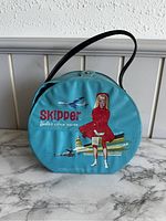 Front view of the round blue Skipper Barbie carrying case showing graphic of Skipper sitting with luggage and airplanes in background.