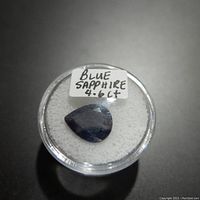 Front view of the pear-shaped blue sapphire gemstone on white foam inside a round clear container with label showing 'BLUE SAPPHIRE 4.6 Ct'.