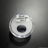 Top view of 4.6 carat oval blue sapphire gemstone in a clear round container with a label displaying 'BLUE SAPPHIRE 4.6 ct'