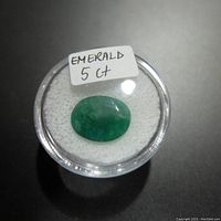 Oval green emerald gemstone approximately 5 carats, shown on white cushion inside a round plastic container. Photo taken for detailed view of stone color and inclusions.