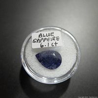 Close-up of pear-shaped blue sapphire gemstone inside round clear plastic display case with white cushion and label indicating 6.1 carats.